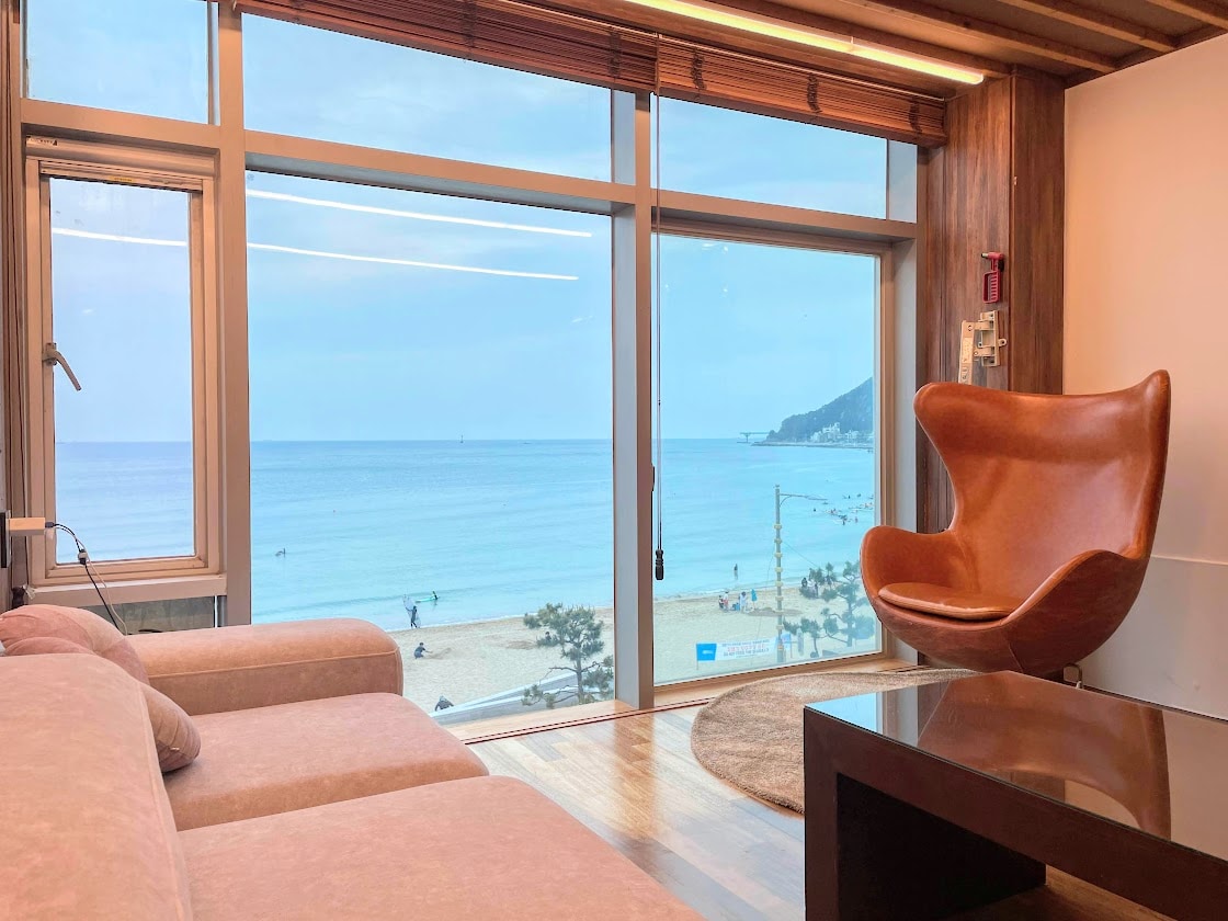 A spacious seating area features a cozy sofa and a sleek coffee table, positioned near large windows that offer a panoramic view of the ocean and beach. The natural light enhances the warm tones of the room, creating a serene atmosphere.