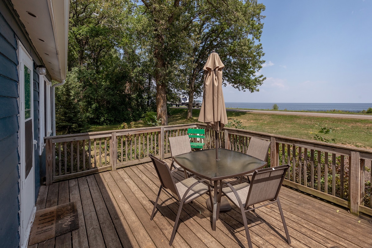 A spacious front deck is equipped with a glass table surrounded by six chairs. A large umbrella provides shade, and a view of the lush greenery and distant lake is visible beyond the railing.