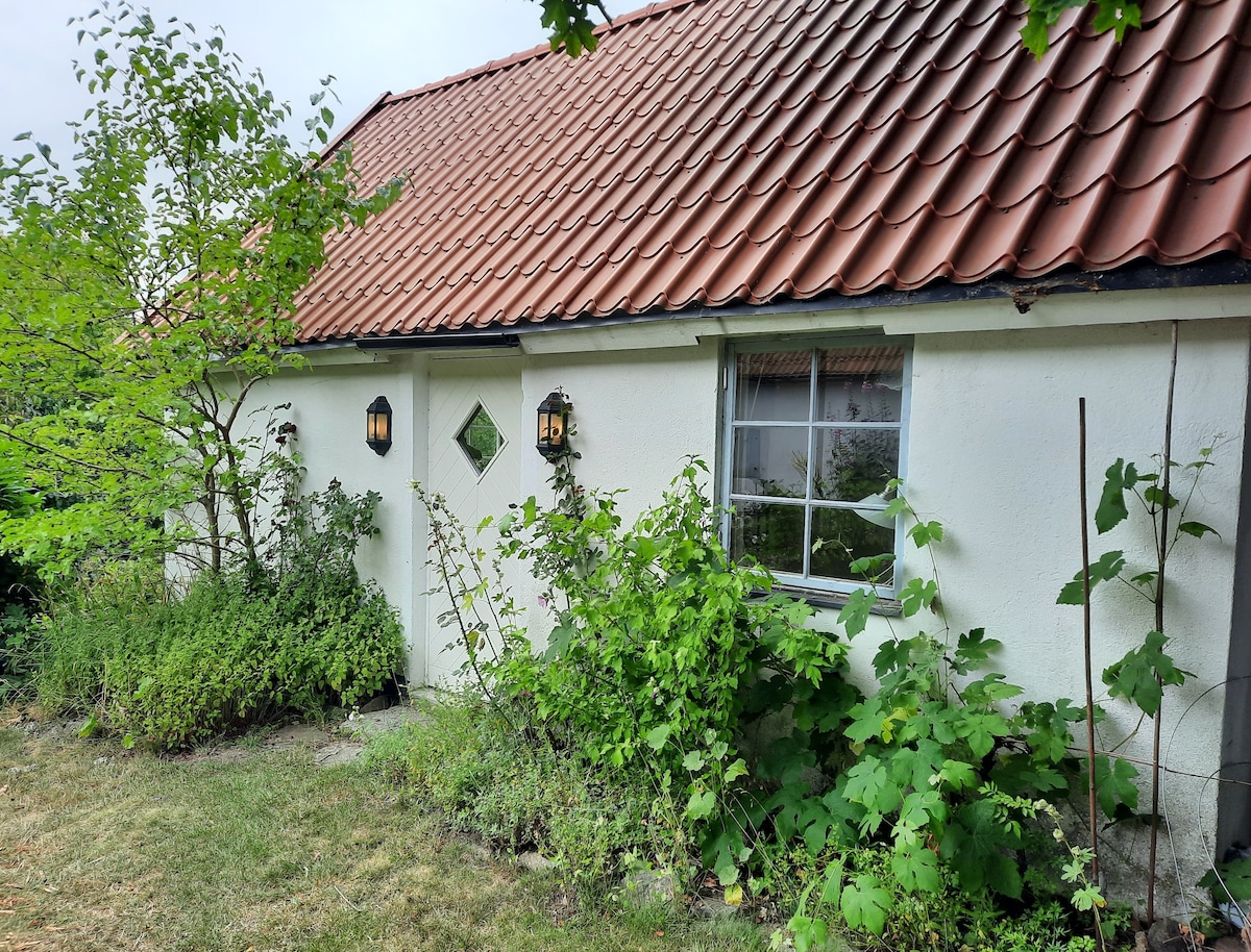 The exterior of the guest cottage is presented, featuring a traditional red-tiled roof and whitewashed walls. Two decorative lanterns are mounted on the façade. Lush greenery and small plants surround the structure, complementing the natural landscape.