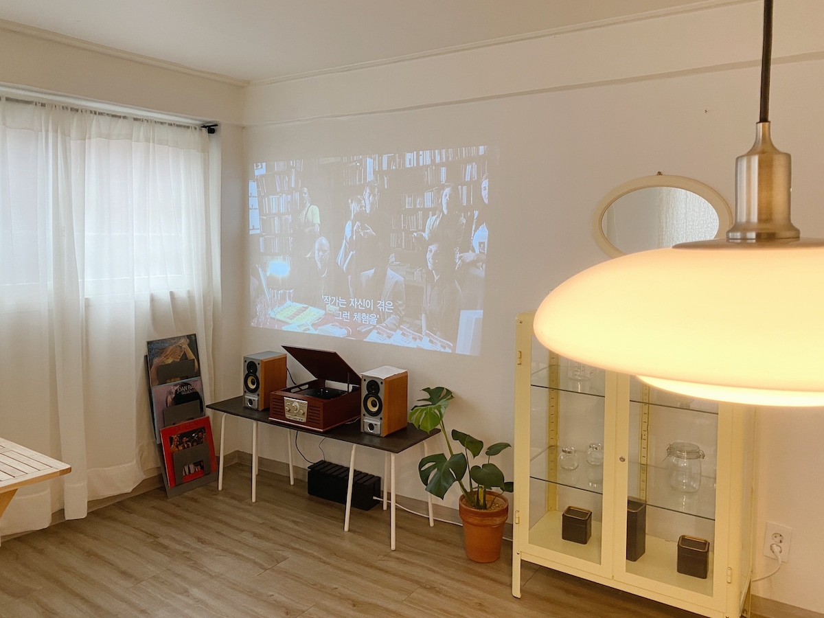 A living area features a projector displaying a film on a white wall, accompanied by a vintage record player and a potted plant. Light filters through sheer curtains, providing a soft ambiance. A cabinet showcases glassware and records, adding a touch of character.