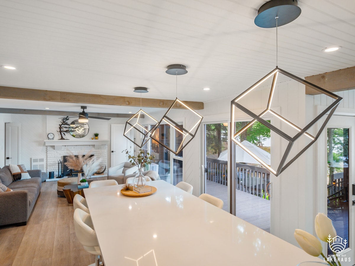 A modern living area features a spacious quartz island with contemporary geometric pendant lights above. A plush sofa is visible in the background, complemented by a well-appointed fireplace. Large sliding glass doors offer views of the deck and outdoor space, enhancing the open layout.
