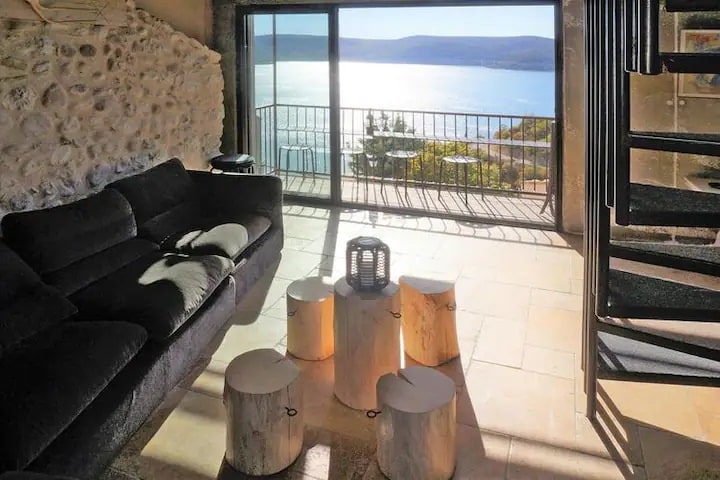 A spacious living area features a dark sofa facing floor-to-ceiling glass doors that open to a scenic view of the water. A rustic wooden coffee table is surrounded by chunky stools. Natural light enhances the inviting atmosphere.