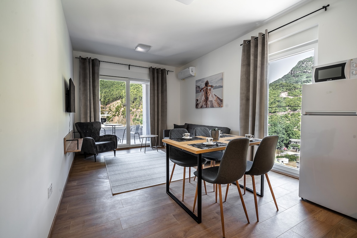 A bright living area with large windows allowing ample natural light. A comfortable sofa and a dining table with four chairs are visible. An air conditioning unit and a flat-screen television are mounted on the wall, complementing the modern furnishings. The balcony is accessible through glass doors.