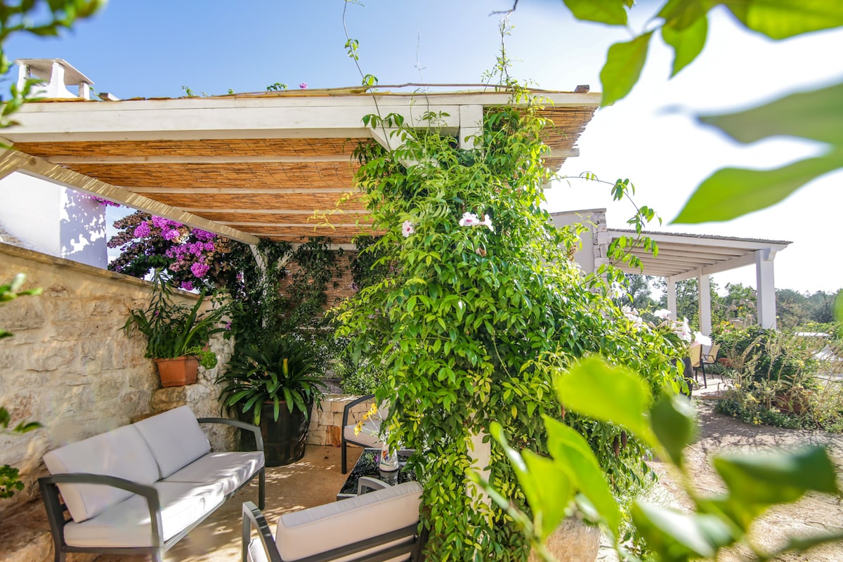 A shaded outdoor seating area is surrounded by lush greenery and flowering plants, providing a serene setting. Comfortable seating, including two chairs and a small table, invites relaxation. A rustic stone wall forms the backdrop, enhancing the natural ambiance of the space.