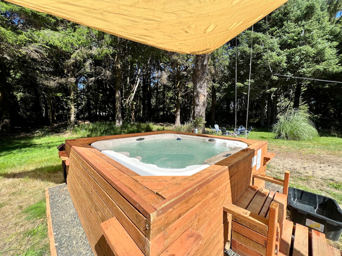 A private hot tub with wood paneling is set in a spacious backyard, surrounded by greenery and tall trees. Shade is provided by a large canopy overhead, and nearby wooden seating is visible, creating an inviting outdoor relaxation space.