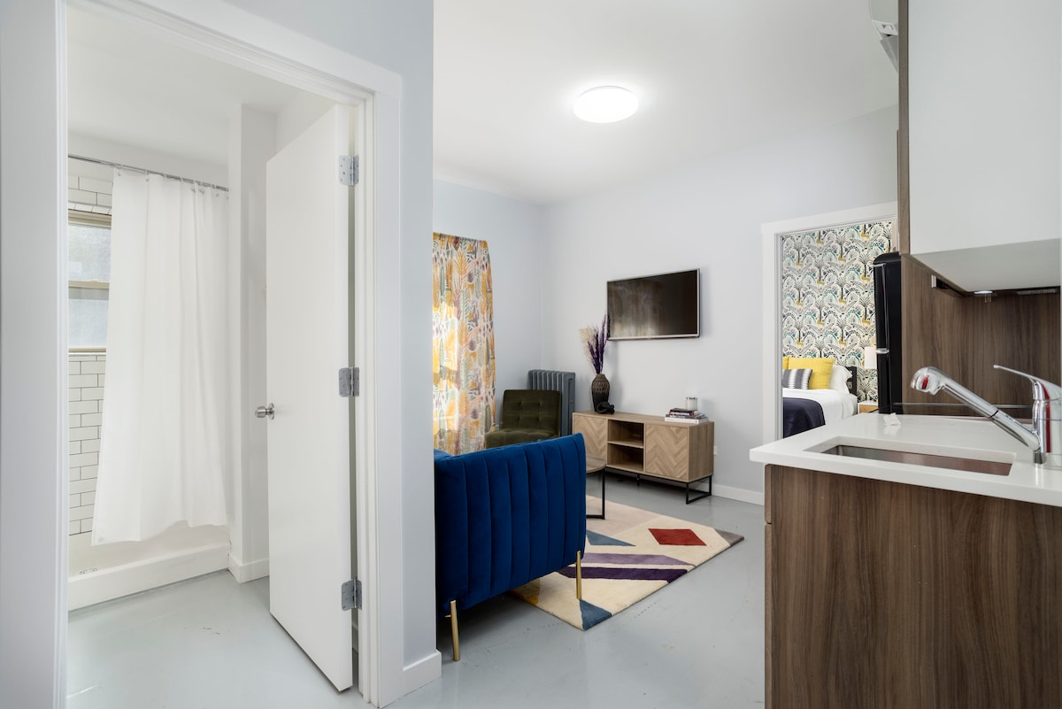 A bright and modern living space is visible, featuring a blue sofa and a flat-screen TV mounted on the wall. The kitchen area is partially shown, showcasing sleek cabinetry. Natural light enters through a window, and a walk-in shower is accessible in the background, surrounded by white curtains.