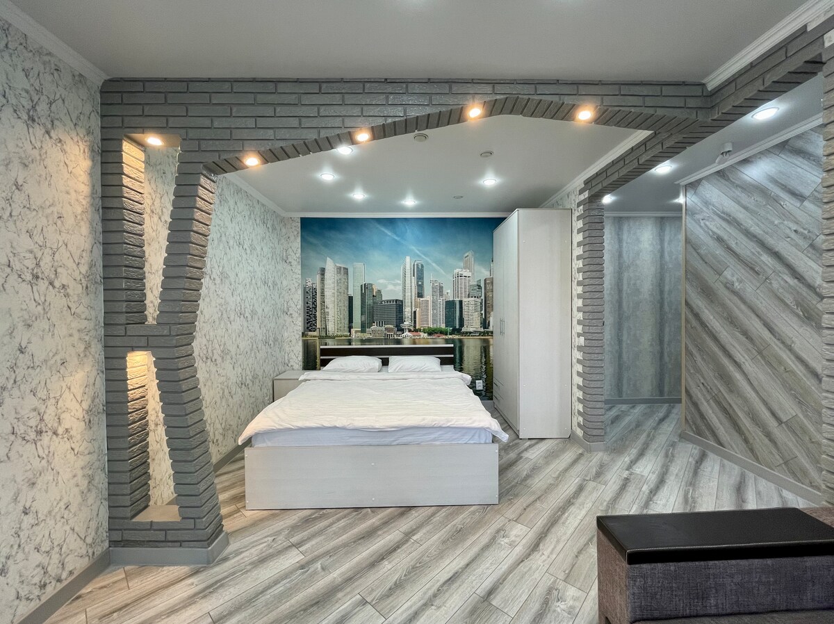 A spacious sleeping area features a large bed against a wall mural of a city skyline. Unique brick accents frame the space, complemented by soft lighting. Light-colored flooring enhances the room's brightness, with modern decor creating a balanced and inviting environment.
