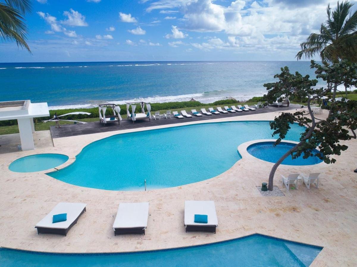 A large outdoor pool features various sections with inviting turquoise waters, surrounded by lounge chairs and cabanas. A beautiful ocean view extends beyond the pool area, framed by palm trees swaying gently in the breeze, creating a serene coastal environment.