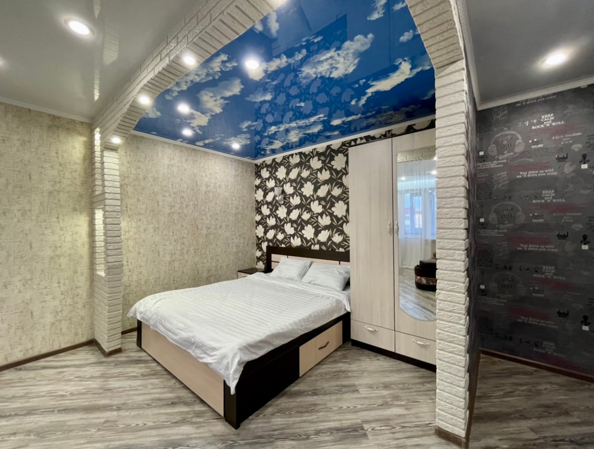 A well-lit studio apartment features a spacious bed with crisp white linens. The ceiling is adorned with a blue sky design, and a stylish wall with dark patterned accents is visible. A white wardrobe adds functional storage, while warm lighting enhances the overall ambiance.