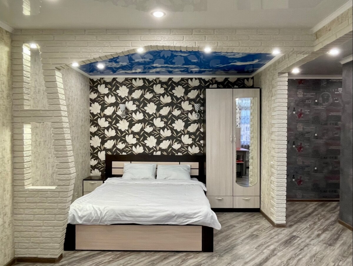 A clean and modern studio features a spacious bed with crisp white linens. A patterned accent wall enhances the design, while a mirrored wardrobe reflects light. Soft overhead lighting creates a bright yet cozy atmosphere, complemented by neutral-toned walls and flooring.