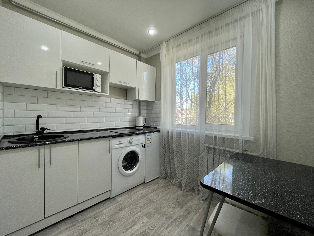 A modern kitchen features glossy white cabinets and a dark countertop. Natural light floods the space through a large window dressed with sheer curtains. Essential appliances, including a microwave and washing machine, are neatly arranged. A small dining table sits in the corner, complementing the overall design.