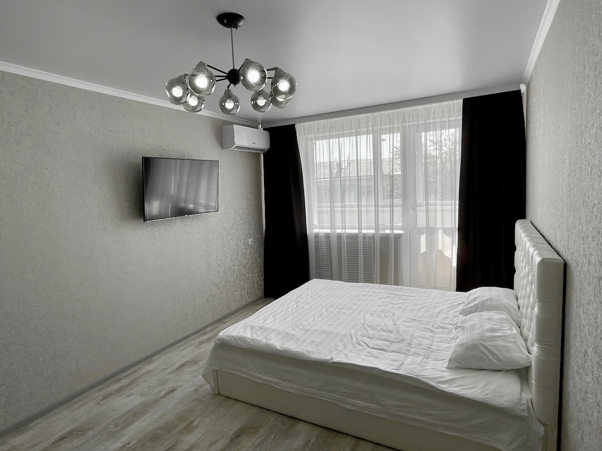 A spacious bedroom features a large bed with a minimalist design, complemented by light-colored bedding. A wall-mounted television is positioned across from the bed. Two large windows allow natural light to fill the room, adorned with sheer and dark curtains for privacy.