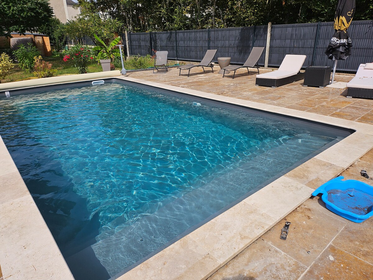 A swimming pool is surrounded by a stone patio, featuring a mix of lounge chairs and a small table. Lush greenery and decorative plants are visible in the background, contributing to a tranquil outdoor setting.