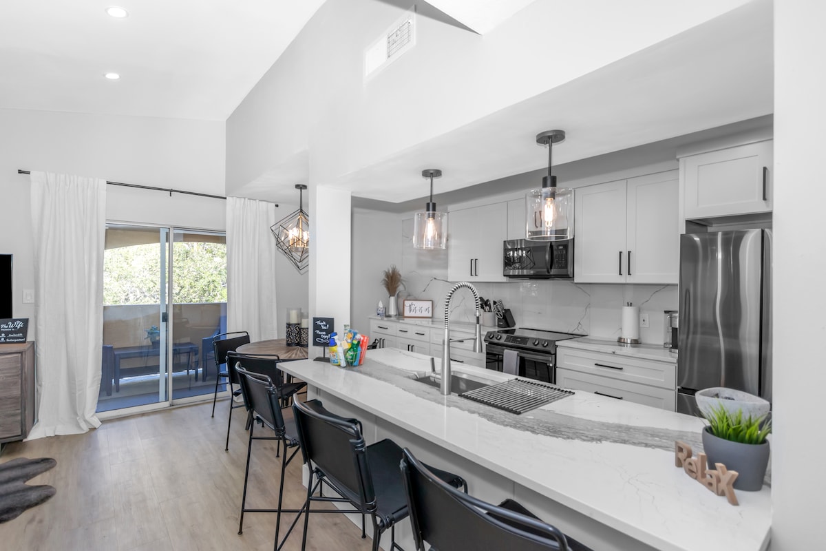 A modern kitchen area is highlighted with sleek white cabinetry and stylish pendant lighting. A large island with seating is arranged alongside stainless steel appliances. Natural light enters from sliding glass doors that open to a scenic outdoor space.