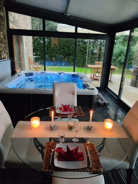 An elegantly set dining table is positioned next to a glass enclosure featuring a hot tub. Soft candlelight enhances the space. A view reveals a well-kept garden just outside, with patio furniture visible in the background.