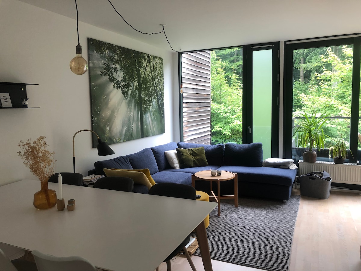 A spacious living area is featured, showcasing a large sectional sofa adorned with green and yellow cushions. A round coffee table sits nearby on a soft rug, while a bright wall art display captures sunlight filtering through trees. Large windows invite views of the surrounding greenery.
