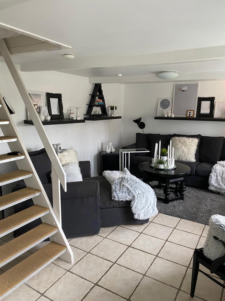 A cozy living area is presented with comfortable seating featuring neutral-colored cushions and a snug throw. A circular table stands at the center, adorned with decorative elements and greenery. A staircase leads to an upper level, while shelves display tasteful decor.