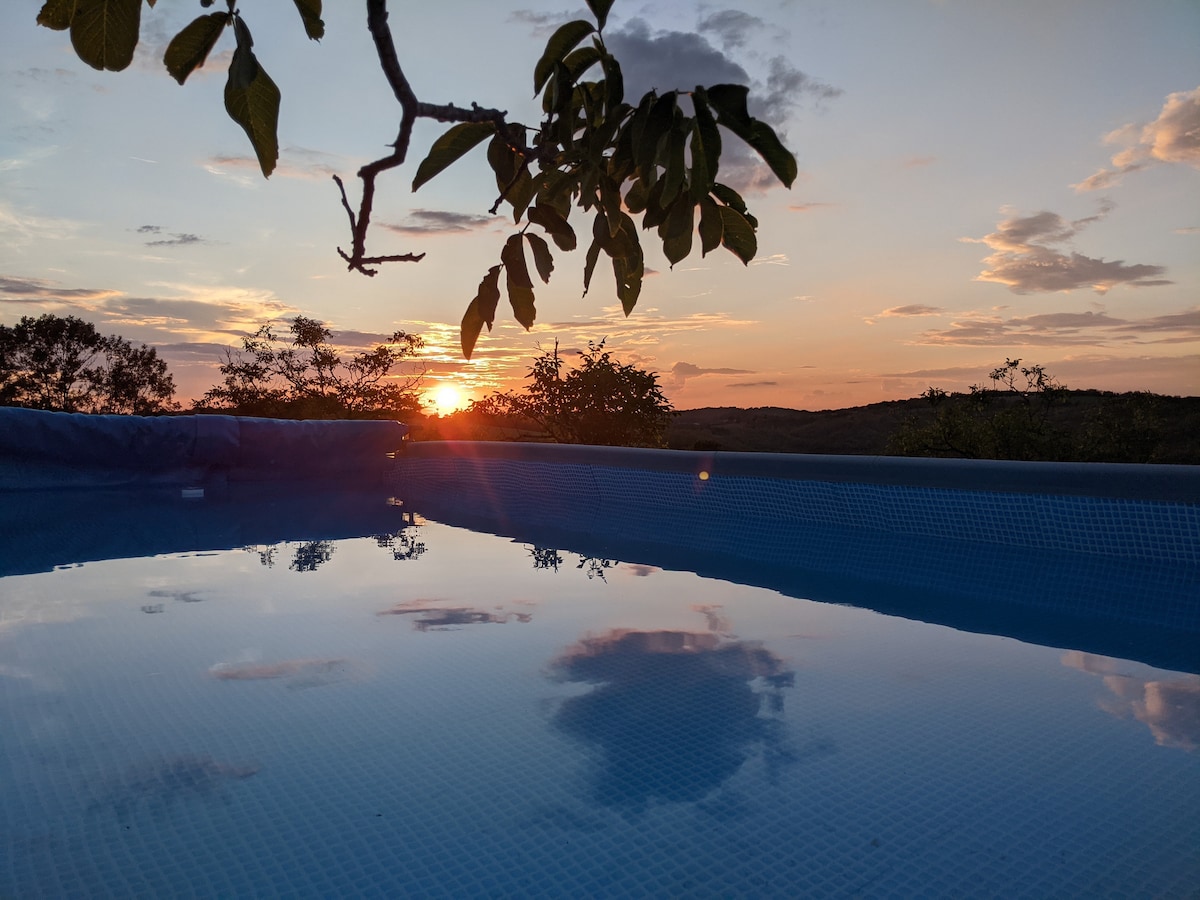 The sunset casts vibrant colors across the sky, reflecting on the calm surface of the swimming pool. The scene is framed by the silhouette of leaves, enhancing the serene atmosphere of the surrounding nature.