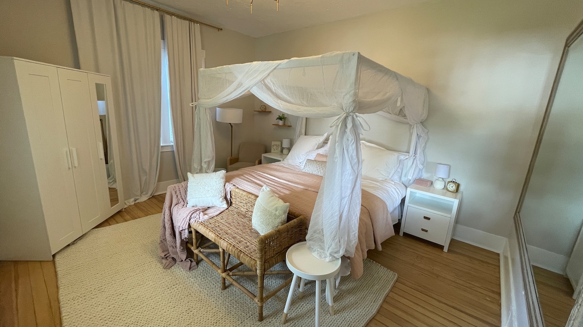 The bedroom features a spacious canopy bed draped with light fabric, complemented by soft bedding and decorative pillows. A seating area with a woven bench and a small round table creates a cozy ambiance. Natural light enters through a large window, illuminating the neutral decor.