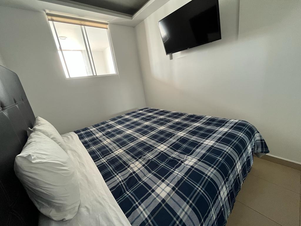A sleeping area features a spacious bed with a plaid blue and white duvet. A flat-screen television is mounted on the wall opposite the bed. Natural light enters through a large window, partially covered by a light shade.