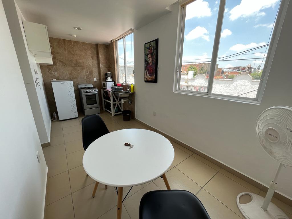 A bright kitchen space features a round white table surrounded by two black chairs. Large windows allow natural light to illuminate the area. A small refrigerator and a stove are visible, alongside minimalist cabinetry, creating a clean and functional environment.