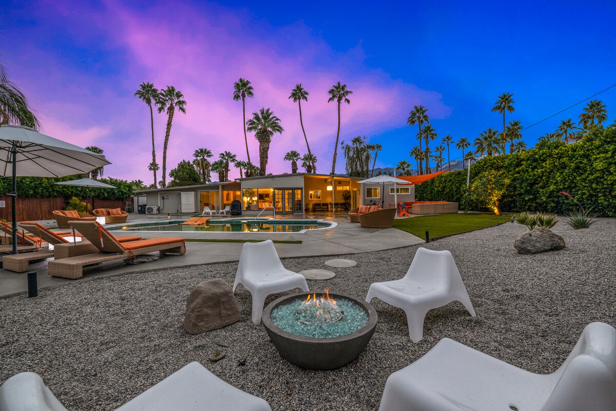 A stylish fire pit is surrounded by modern seating, set against a backdrop of a private backyard with a sparkling pool. Palm trees line the space, with warm evening skies transitioning from purple to blue, creating a serene atmosphere for outdoor gatherings.