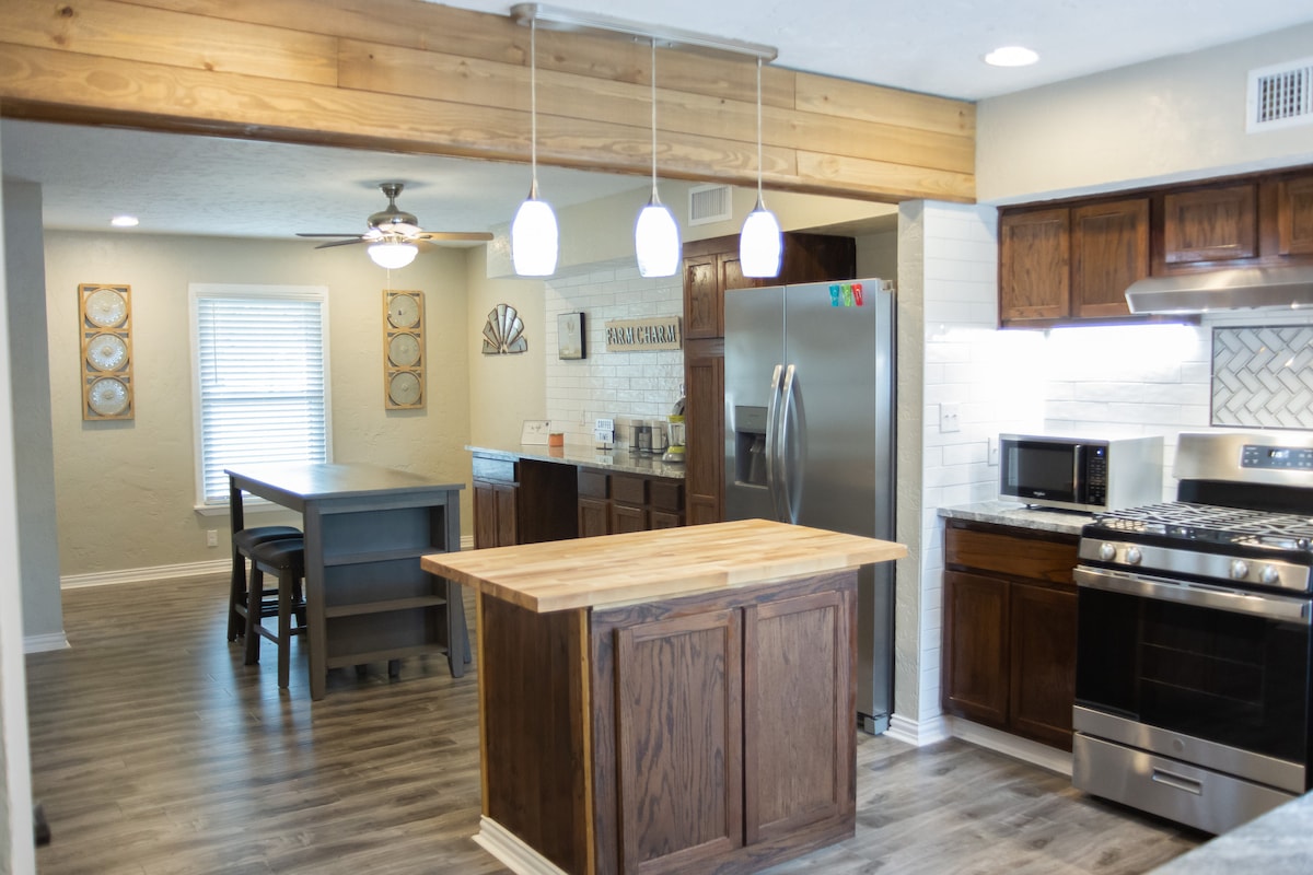 A modern kitchen features dark wooden cabinetry and a large central island with a natural wood countertop. Stainless steel appliances and a microwave are visible. Pendant lights hang above, providing illumination. A dining table is placed near a window, adding a functional dining space.