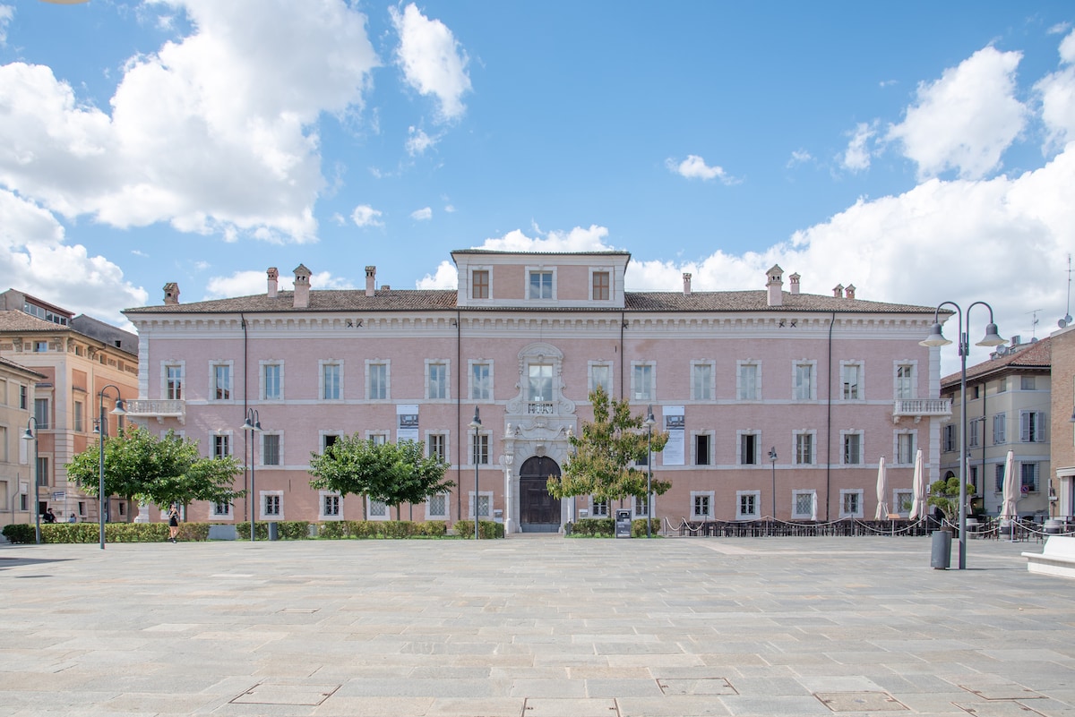 A historical building is displayed in a central square, with a pink façade and classic architectural details. The entrance features large windows and a central archway, framed by greenery. Bright clouds fill the blue sky, enhancing the inviting atmosphere of the surrounding plaza.