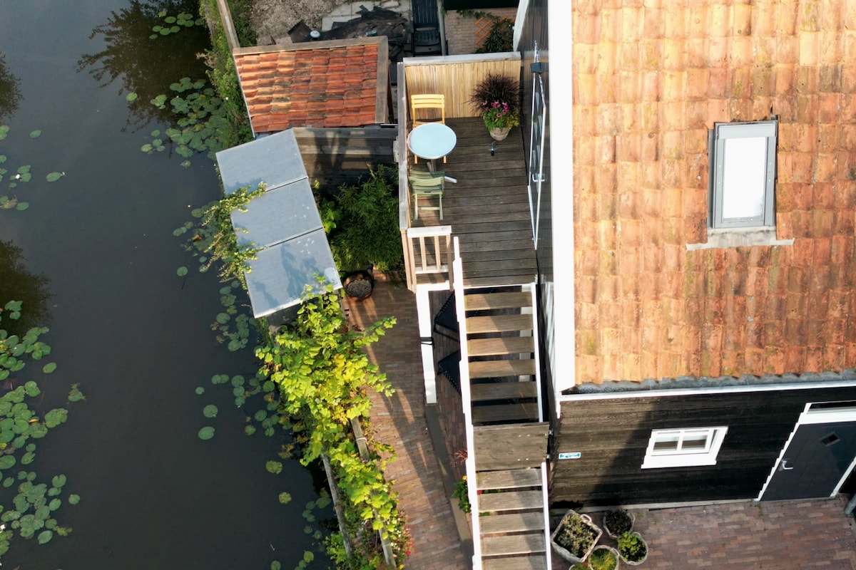 An aerial view reveals a wooden walkway leading to the water, with a small table and two chairs on a terrace. The staircase provides access to the first floor. Lush greenery and potted plants are positioned near the edges, adding a natural touch to the surroundings.