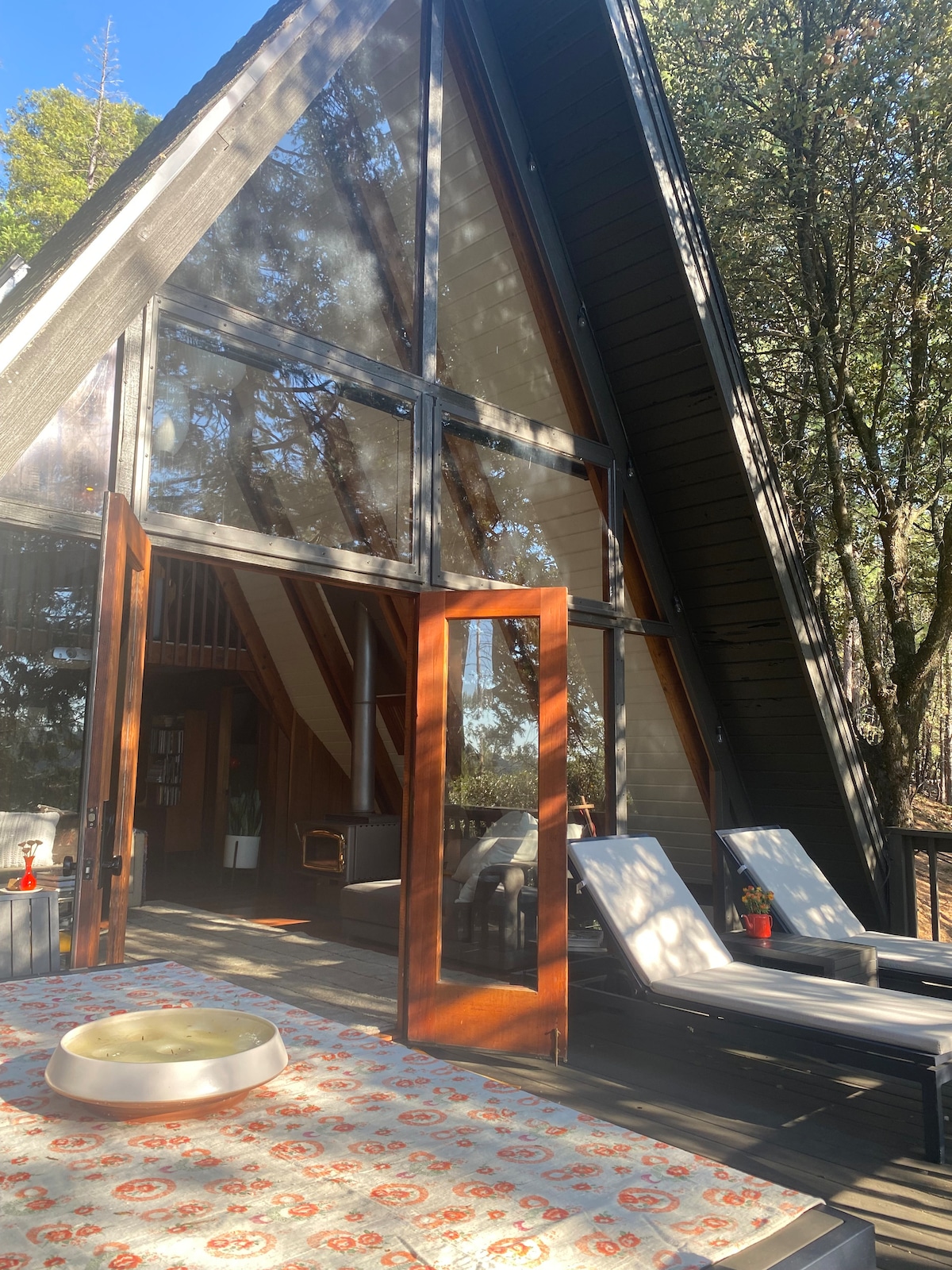 The A-frame cabin features large glass panels that bring in natural light, framed by wooden doors that open to a spacious deck. Two lounge chairs are positioned on the deck, with a small table adorned with a decorative bowl at the forefront.