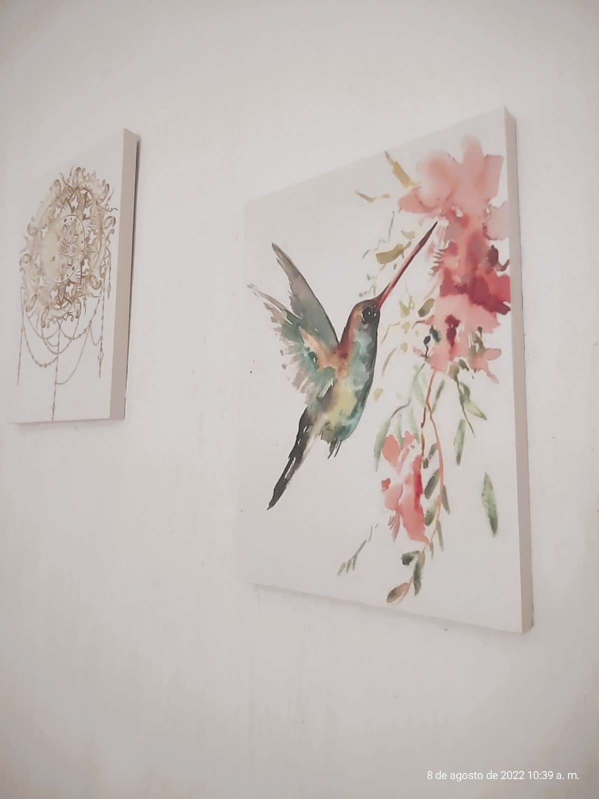 Two watercolor paintings adorn a light-colored wall. One depicts a vibrant hummingbird with pink flowers, while the other features an intricate golden chandelier. The artwork adds a touch of color and detail to the space.