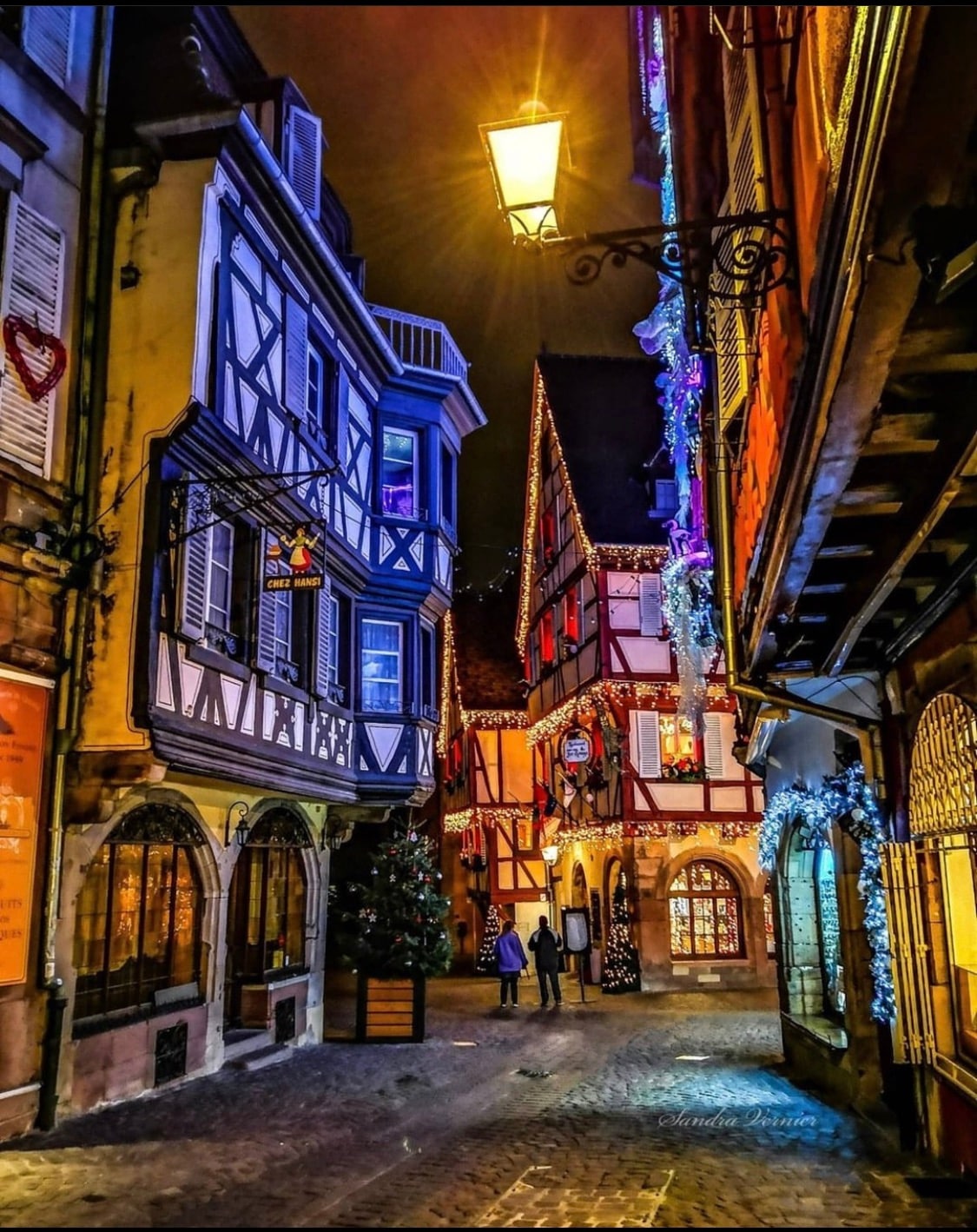 A charming narrow street is illuminated by warm street lamps and festive decorations. Half-timbered buildings with colorful facades line both sides, creating a cozy atmosphere. Joyful figures can be seen walking, while a decorated Christmas tree adds to the festive ambiance, enhancing the scene's seasonal appeal.