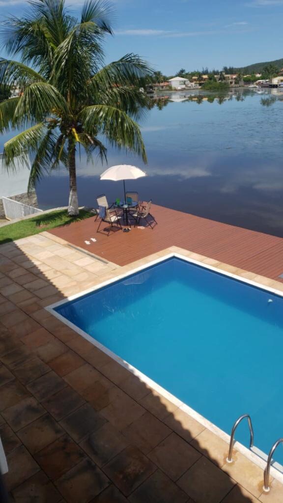 A clear, inviting swimming pool is framed by a stone patio, with pool steps visible. Nearby, a wooden deck features a round table and two chairs, shaded by a large umbrella. Tall palm trees add a tropical touch to the scene, overlooking a calm waterway.