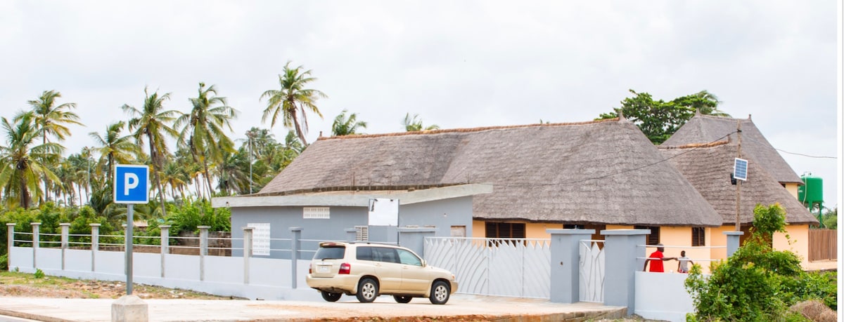 A bungalow with a grass-thatch roof is set within a landscaped area, surrounded by palm trees. A fenced parking area is visible in the foreground, along with an indication for parking. The structure features multiple entrances and a mix of painted walls.