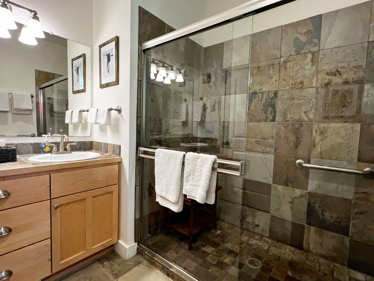 An inviting bathroom features a spacious glass shower with a tiled interior. The wood vanity is complemented by a large circular sink and modern fixtures. Soft towels are neatly arranged on towel bars, with artwork displayed on the walls, adding a touch of style to the space.