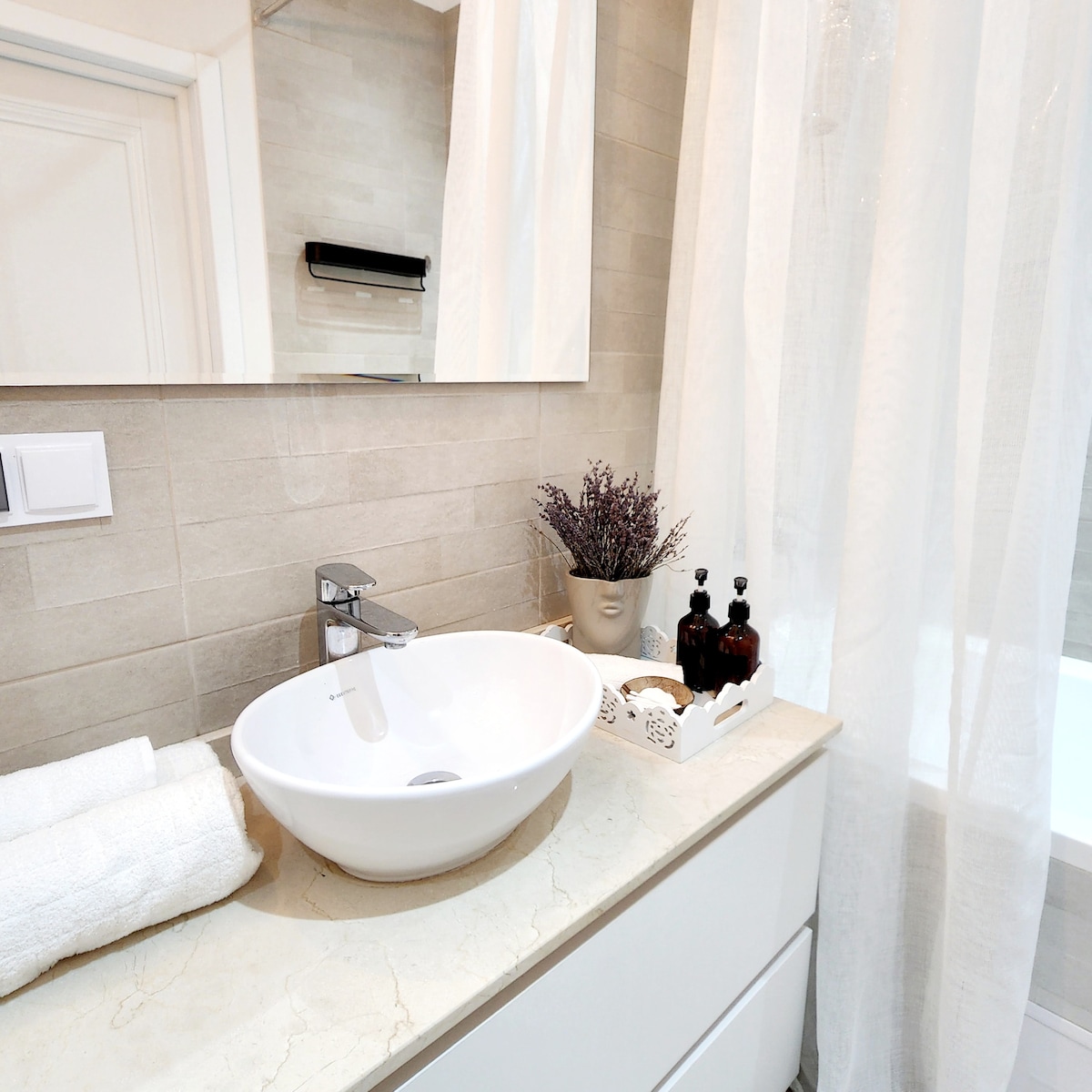 The bathroom features a modern sink with a rounded basin set on a smooth countertop. Soft towels are neatly arranged beside the sink. A decorative pot of lavender and two dark glass bottles are placed on a tray, enhancing the space's tranquility. A light curtain provides privacy.