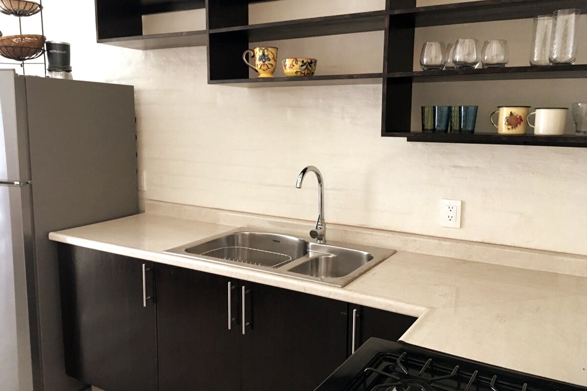 The kitchen area features modern cabinetry with open shelving displaying various colorful dishes and glassware. A sleek countertop with a double sink is positioned beside a stainless steel refrigerator and a gas stove, providing a functional cooking space.