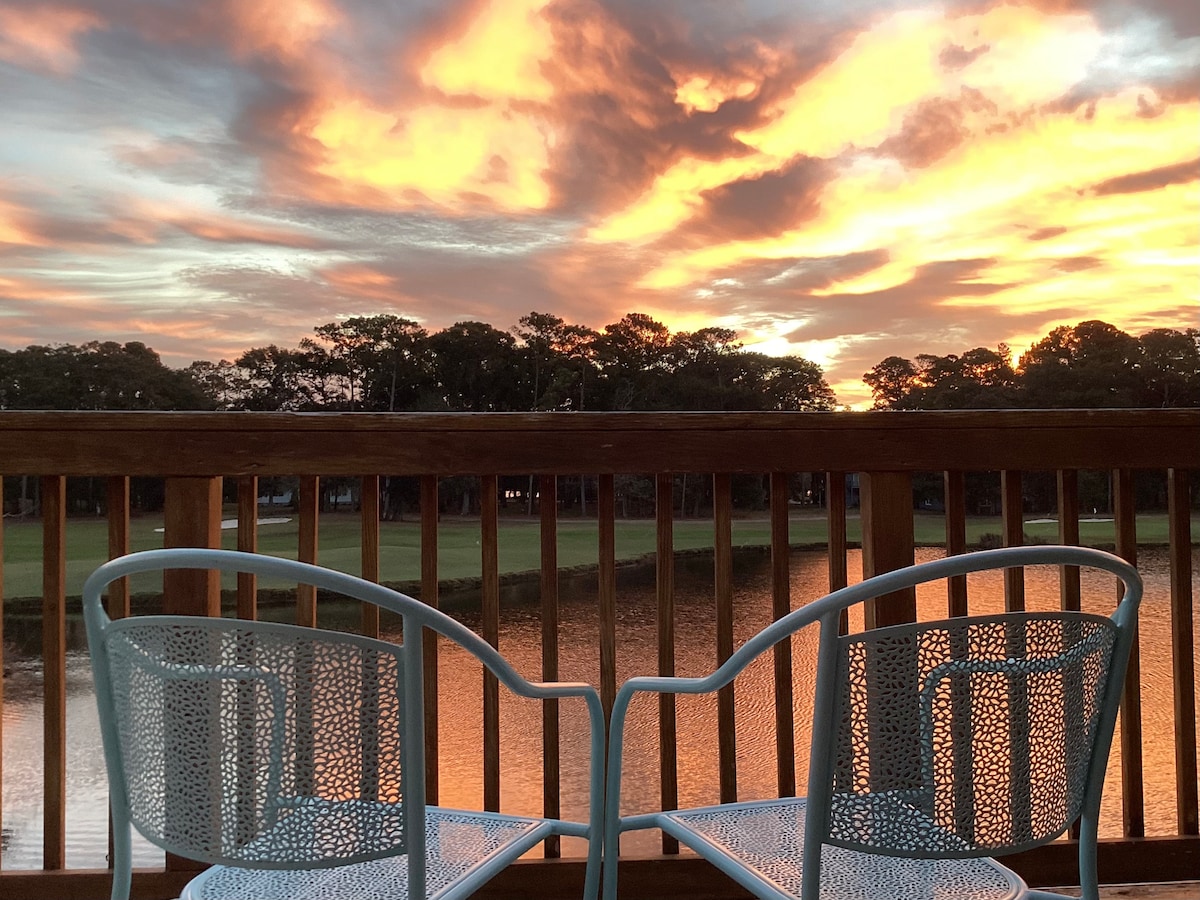 Two light blue chairs are positioned on a deck, facing a stunning sunset that casts warm colors across the sky. Reflected in the water below, the vibrant hues enhance the serene view of the golf course and surrounding trees.