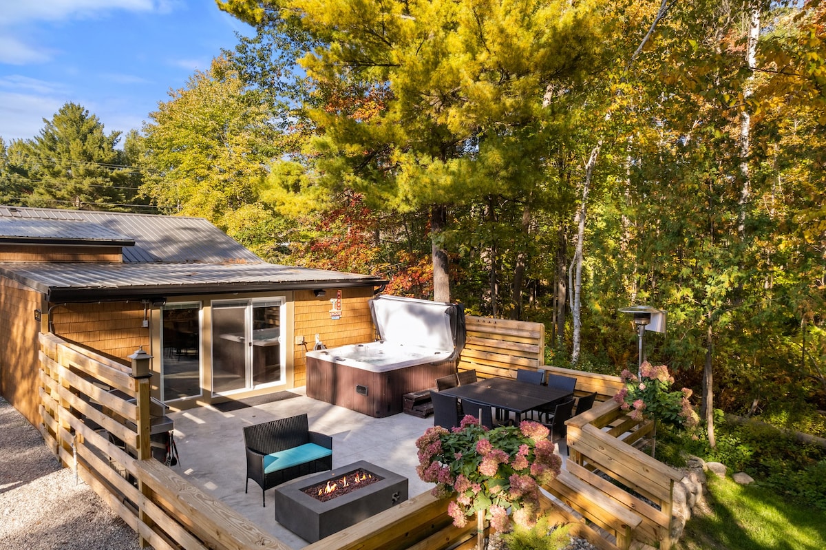 An outdoor patio area features a hot tub, surrounded by wood paneling and lush greenery. A fire pit is present, along with seating options including black chairs and a dining table. The space is designed for relaxation, amid a backdrop of vibrant fall foliage.