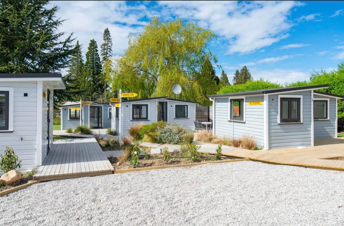 Four renovated units are positioned around a landscaped area, connected by a boardwalk path. The exterior features a combination of wood and light-colored finishes. Soft green shrubbery and gravel surround the units, with tall trees and a bright blue sky in the background.