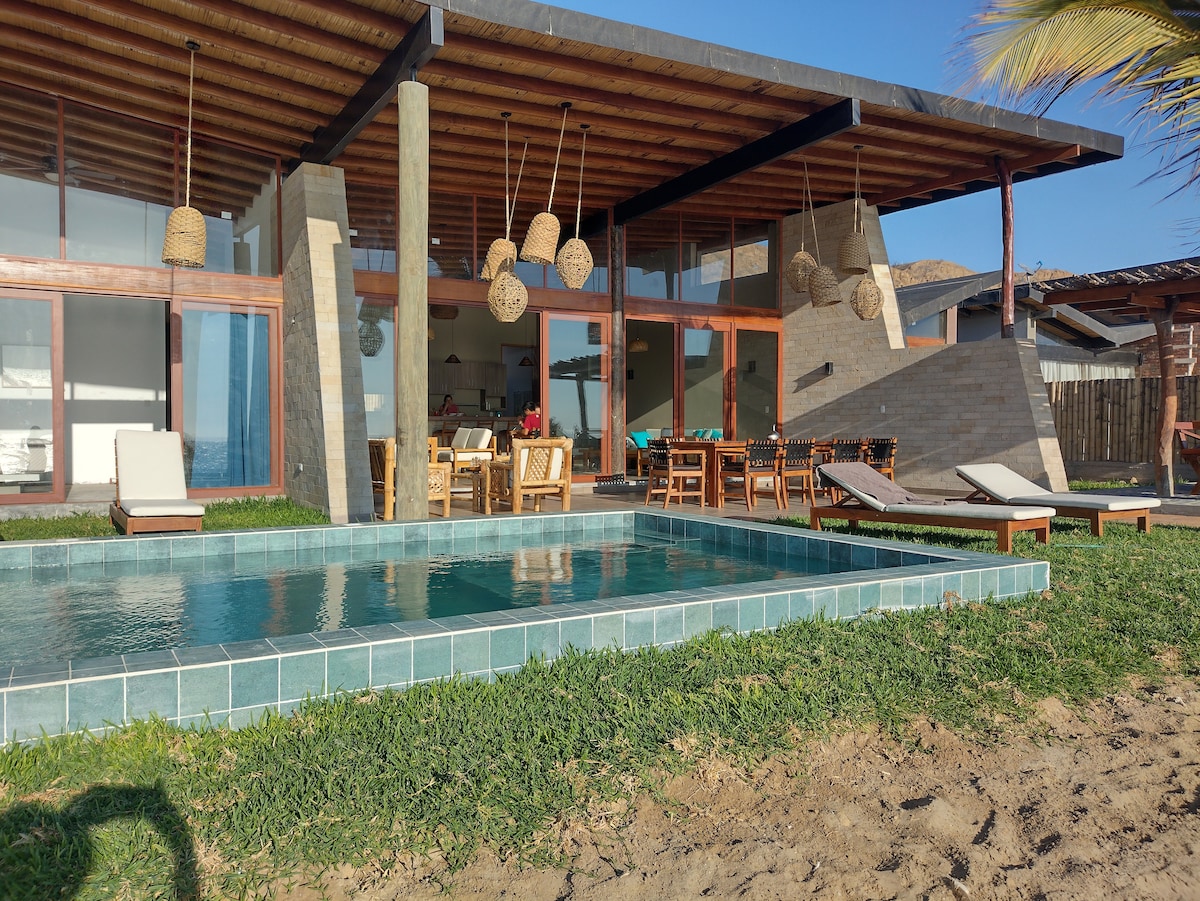 A modern home is showcased with a spacious outdoor area, featuring a private pool bordered by green grass. Comfortable sun loungers are arranged alongside, while a dining area and shaded seating can be seen beneath the wooden overhang.