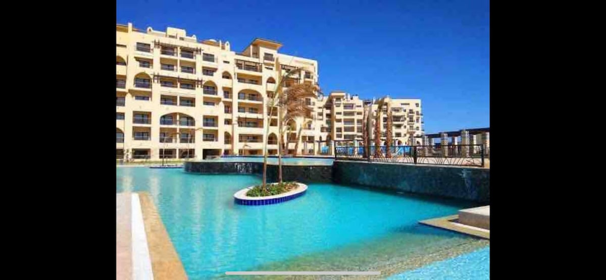 A large swimming pool is showcased, featuring clear turquoise water and a central island with palm trees. Surrounding structures exhibit modern architecture with arched balconies. The bright blue sky adds to the serene ambiance of the scene.