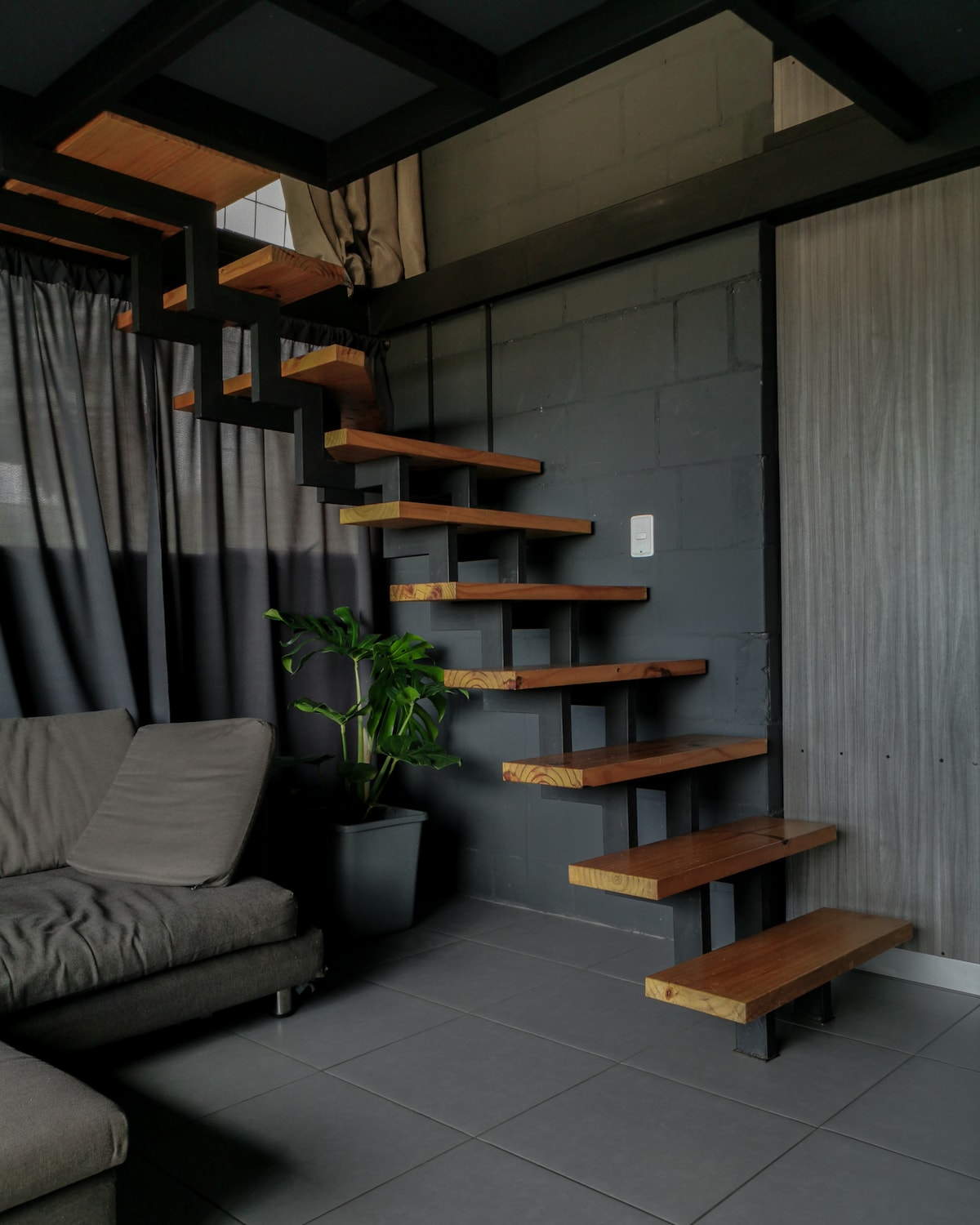 A modern staircase with wooden steps is seen ascending alongside a gray wall. A leafy green plant in a black pot is positioned near a comfortable gray sofa, and dark curtains provide a contrast against the light flooring.