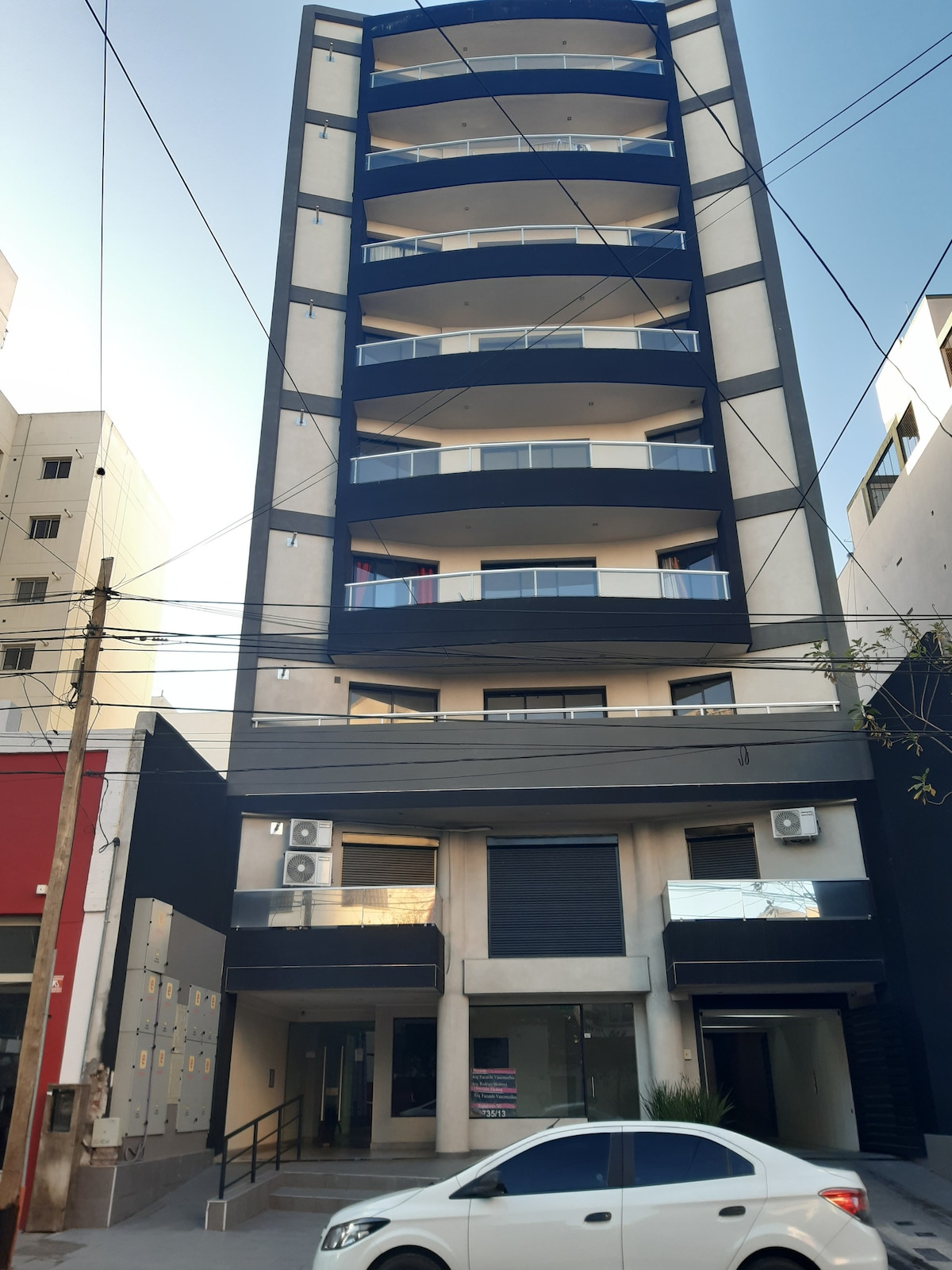 The exterior of a modern multi-story building features a sleek design with balconies on each floor. Large windows provide natural light, while air conditioning units are visible. A parked car is located in front of the building, and the surrounding area includes utility lines and other structures.