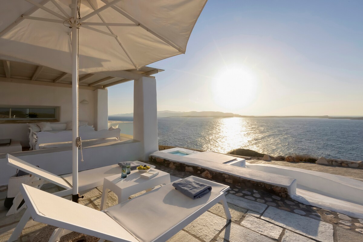 A sunlit outdoor terrace features white lounge chairs beneath a large umbrella. The sea view highlights the shimmering water and sunset. The area is surrounded by a stone wall, providing a serene atmosphere for relaxation.