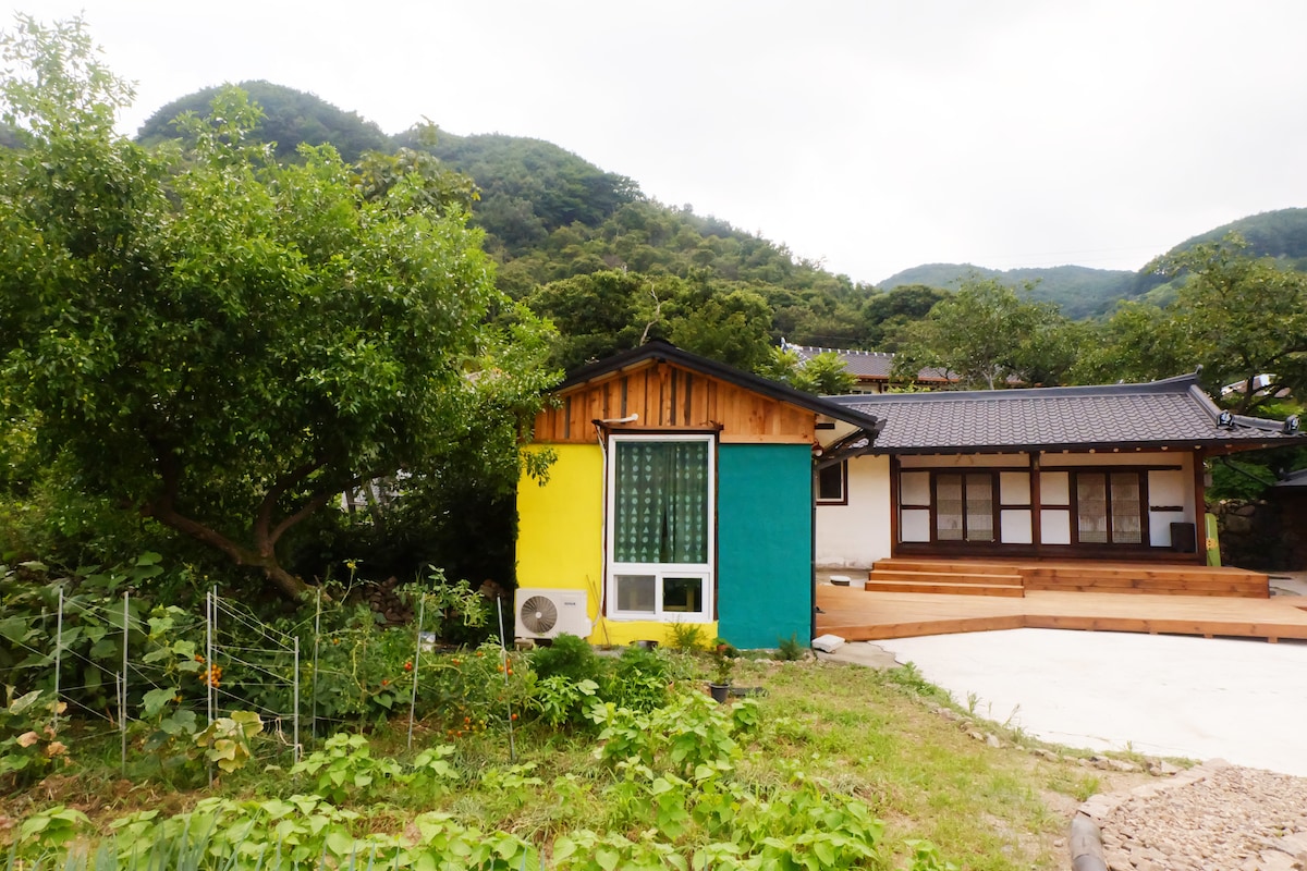 A traditional hanok displays vibrant colors, with a green and yellow façade contrasting against a natural backdrop of hills. A wooden deck extends from the main house, with a garden area visible in the foreground. Air conditioning unit is mounted on the side wall.