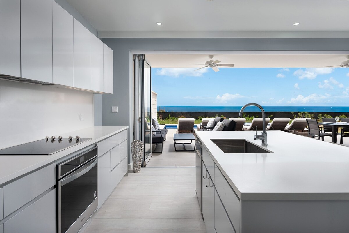 The modern kitchen features sleek white cabinetry and a spacious island with a sink. Large glass doors provide a view of the outdoor patio, showcasing seating and sun loungers with the sea visible in the background.