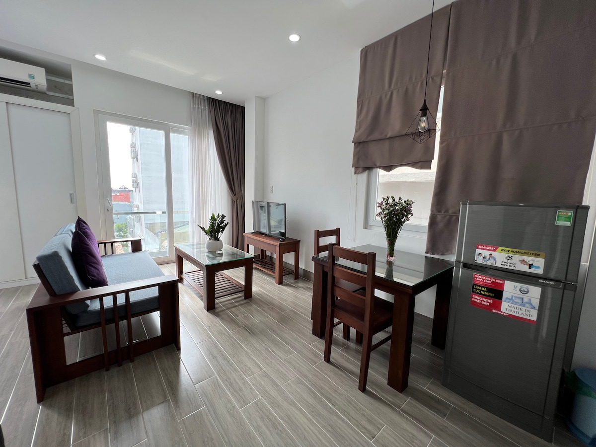 A bright living area features a blue sofa against the wall, with a glass dining table and wooden chairs nearby. A television sits on a wooden stand across from the seating. Large windows allow natural light to fill the space, complemented by neutral-colored curtains.