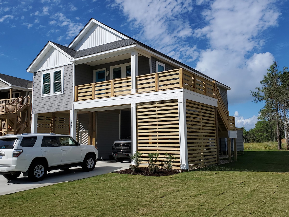 A two-story house is showcased with a modern exterior, featuring grey siding and white accents. The elevated porch is accessible by a staircase, and a carport is visible beneath. Lush green grass surrounds the structure, complemented by a sunny blue sky dotted with clouds.