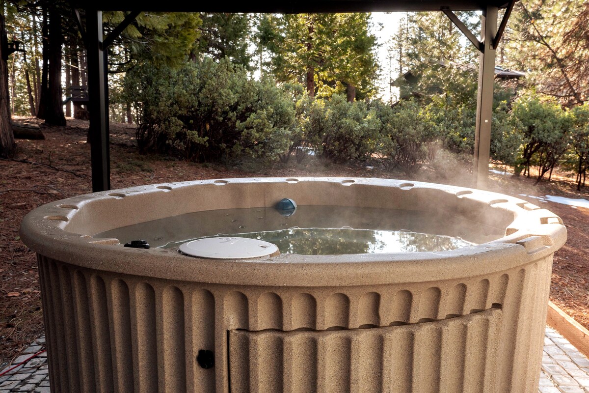 A large, seven-person hot tub is situated under a shaded gazebo surrounded by tall trees. Steaming water reflects the natural light, creating a serene ambiance. The textured exterior of the hot tub contrasts with the calm landscape of greenery and pine needles on the ground.